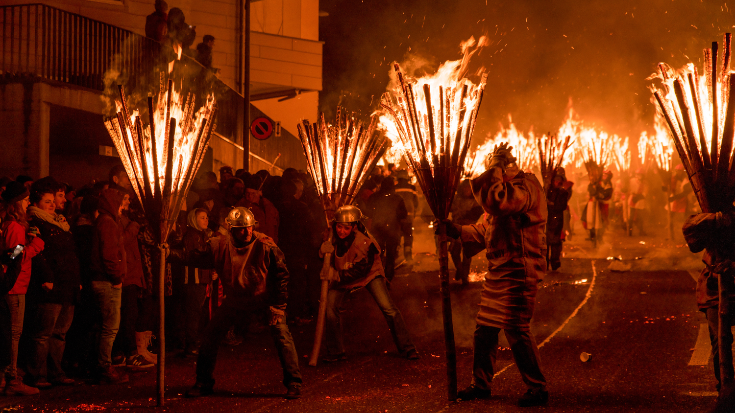 Des personnes portant de grands faisceaux de branches enflammés défilent dans une rue bondée la nuit, entourées de spectateurs, lors d’un événement traditionnel.