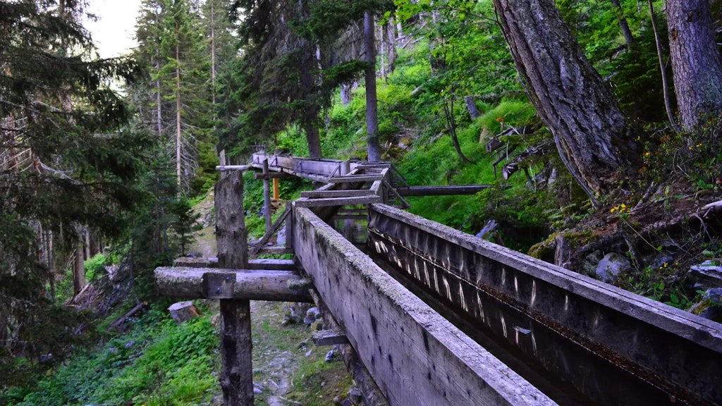 Canal d’irrigation en bois traversant une forêt de montagne, soutenu par des structures en bois, avec un sentier et des arbres autour.