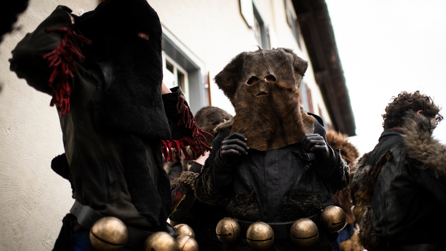 Des personnes en costumes noirs avec des masques en fourrure et des grelots attachés à leur ceinture participent à un événement traditionnel dans une rue de village.