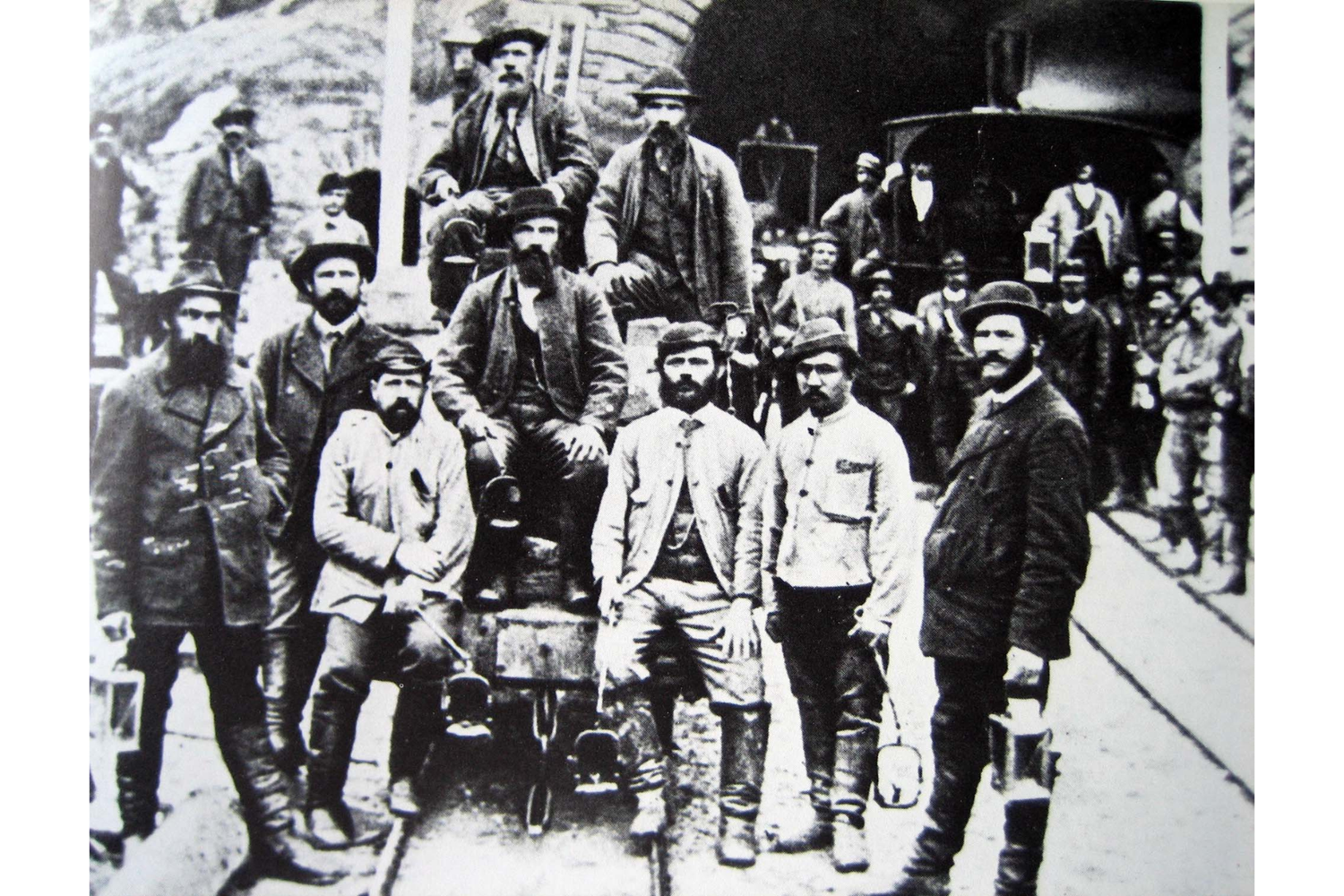 Groupe d’ouvriers du tunnel du Saint-Gothard à Airolo vers 1880, posant en costume de travail devant l’entrée du chantier, outils et wagonnets autour d’eux.