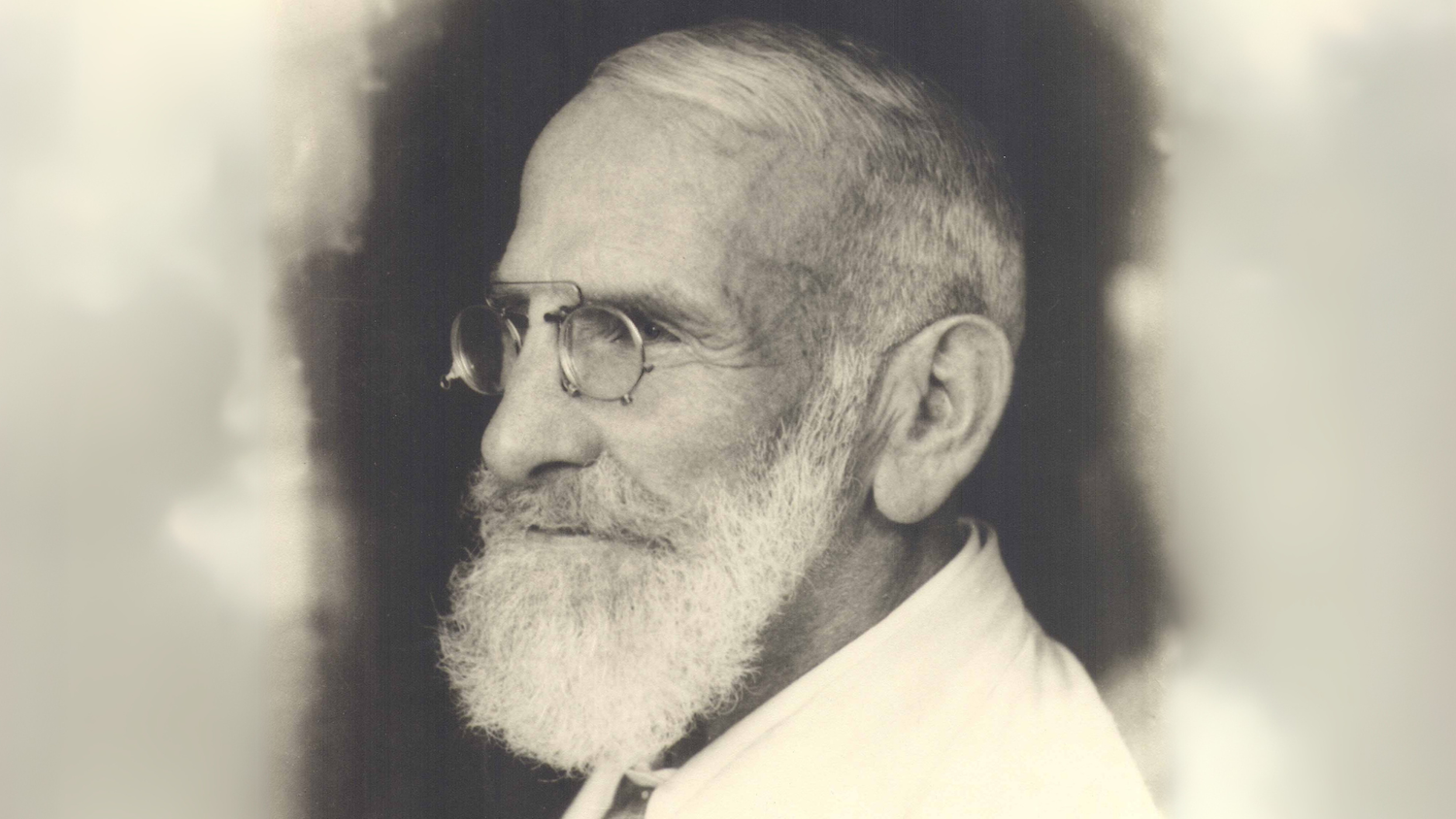 Portrait en noir et blanc d’un homme âgé avec une barbe blanche, des lunettes rondes et un costume clair, vu de profil sur un fond flou.