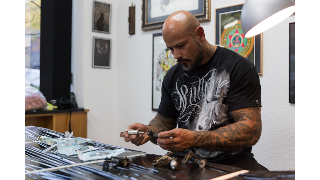 Photographie montrant un homme tatoué assis dans un atelier, manipulant du matériel de tatouage sur une table, avec des cadres accrochés au mur derrière lui.