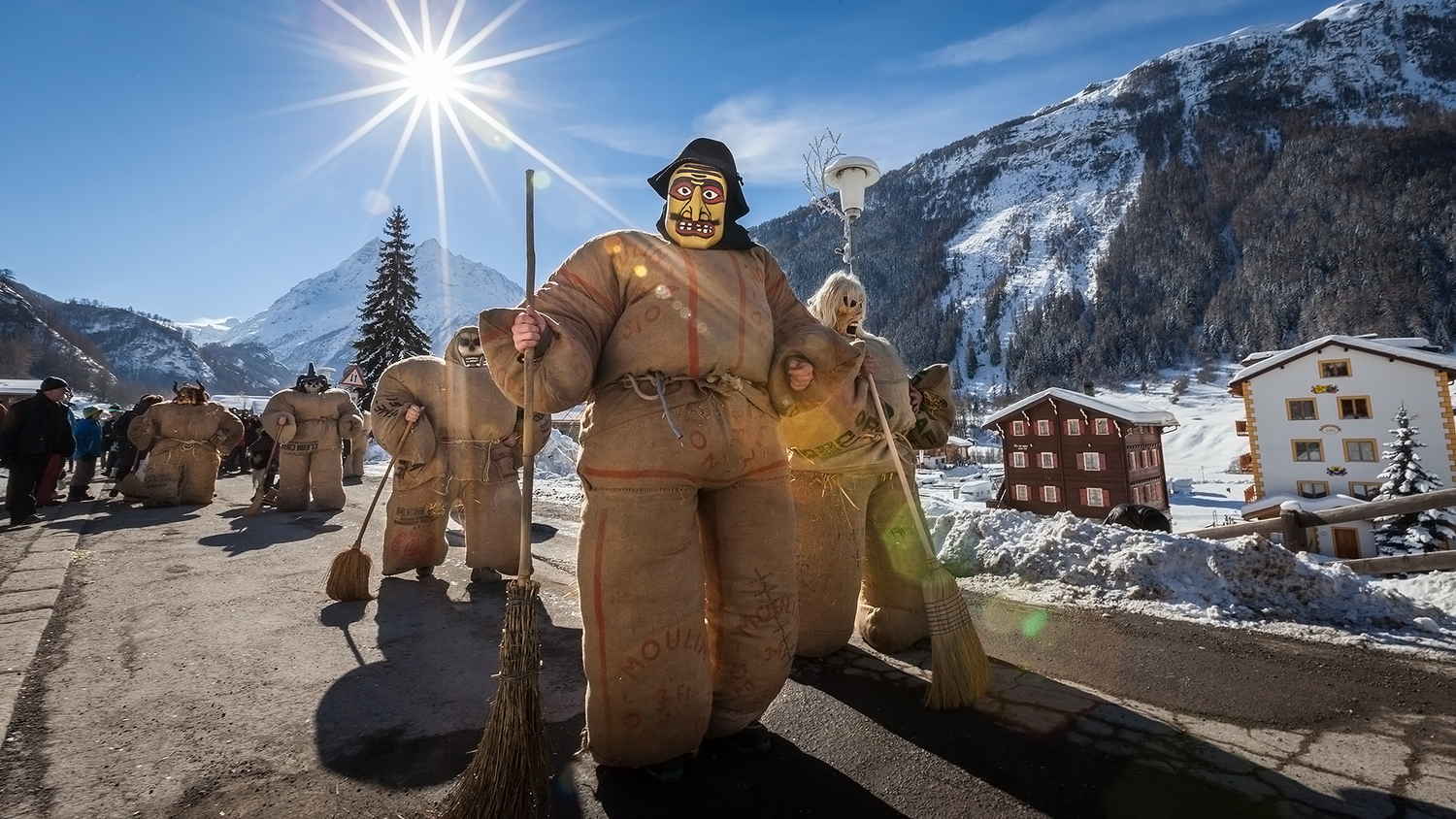 Des personnes en costumes rembourrés de foin avec des masques colorés et des balais défilent dans une rue enneigée d’un village alpin sous un ciel ensoleillé.