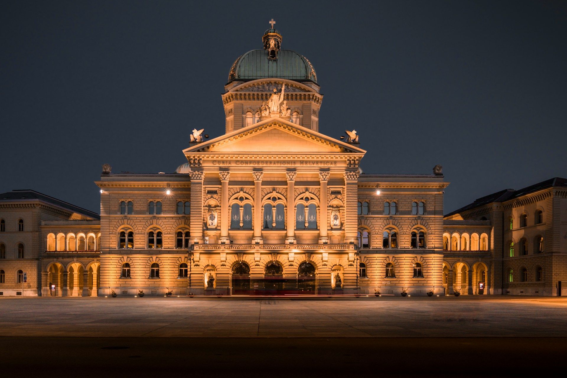 Vue de nuit du Palais fédéral suisse à Berne, éclairé, présentant sa façade symétrique en pierre avec arcades, colonnes et coupole, devant une vaste esplanade vide. 