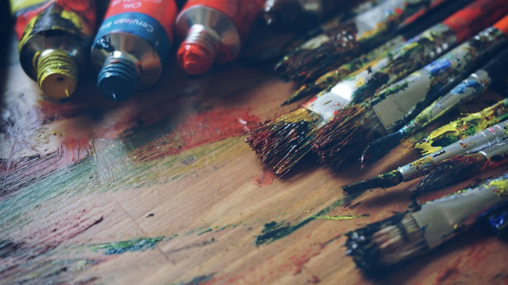 Plusieurs tubes de peinture à l’huile ouverts et des pinceaux tachés de peinture posés sur un support en bois couvert de traces colorées.