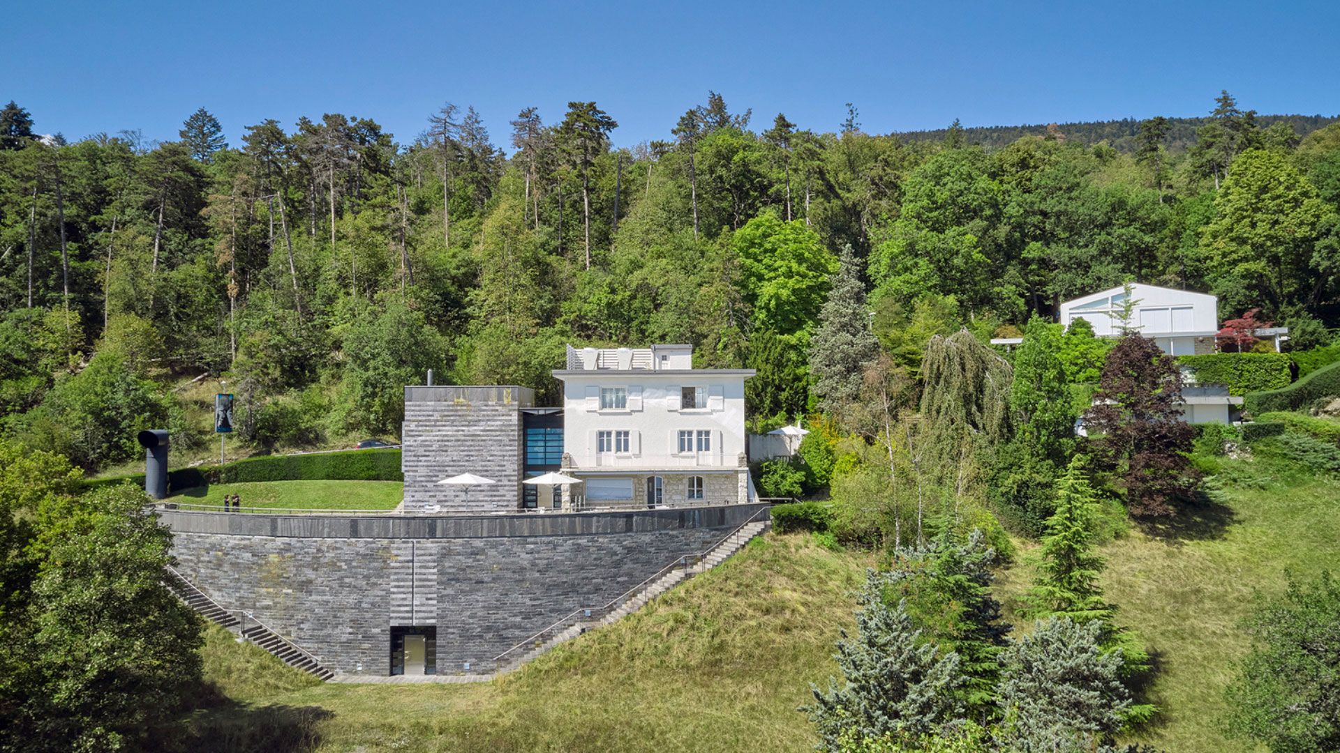 Vue du Centre Dürrenmatt Neuchâtel avec bâtiment blanc et extension en pierre grise, entouré de végétation et situé au pied d’une colline boisée.