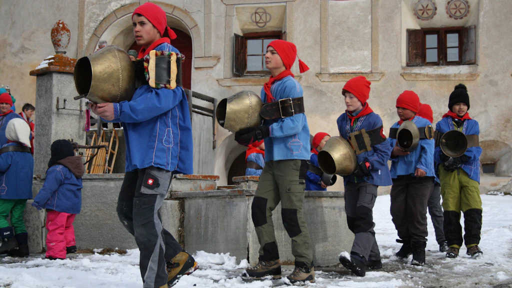 Des enfants en chemises traditionnelles bleues et bonnets rouges passent dans la neige avec de grandes cloches de vache près d’une fontaine de village.