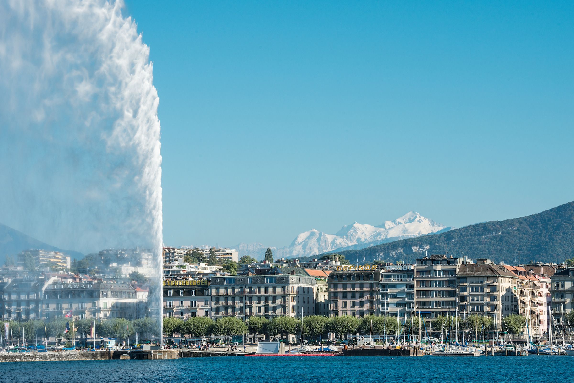 Jet d’Eau dominant le lac à gauche, front de bâtiments au bord de l’eau, montagnes enneigées en arrière-plan et ciel bleu uni.