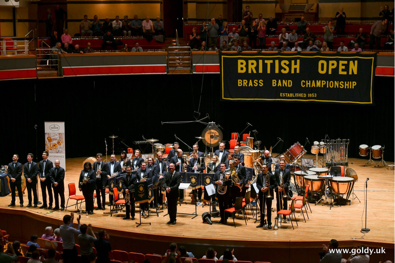 Photo d’un ensemble de cuivres en smoking aligné sur scène devant un fond noir, instruments et pupitres disposés sous une banderole «BRITISH OPEN Brass Band Championship», public en gradins.