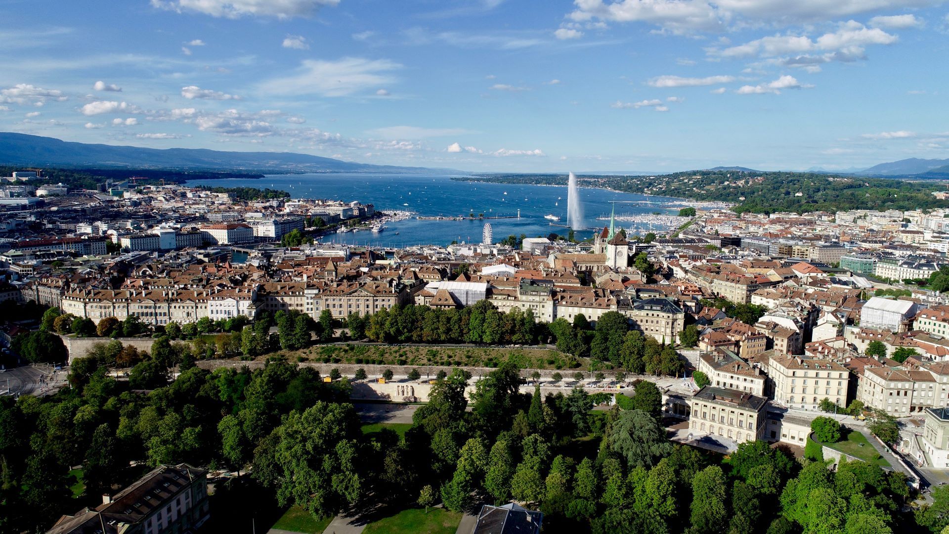 Vue plongeante d’une ville au bord d’un lac, Jet d’Eau visible sur le lac, toits en tuiles rouges, arbres en bas de l’image et collines à l’horizon.