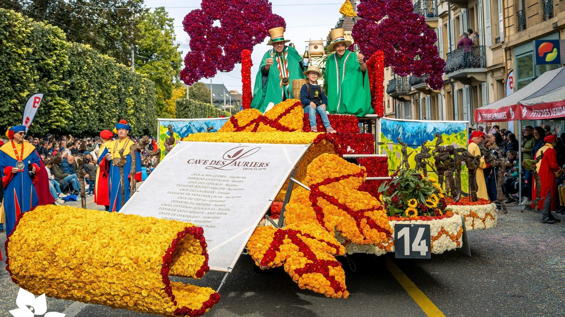 Char fleuri de la Cave des Auriers avec deux adultes en vert et un enfant, décoré de fleurs colorées et de feuilles, défilant à la fête des vendanges de Neuchâtel.