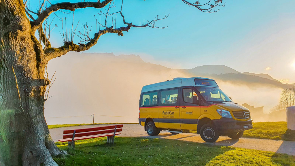 Minibus jaune PubliCar garé près d’un banc rouge au bord d’un chemin, devant un paysage de montagnes et de brume au lever du soleil, arbre dénudé à gauche.