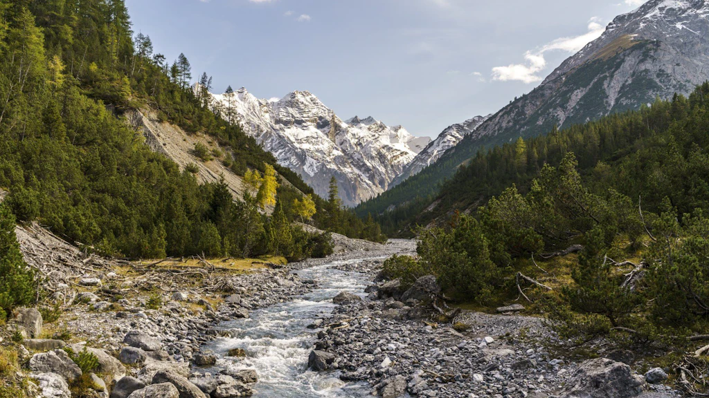 Un ruisseau de montagne serpente au fond d’une vallée rocailleuse, entourée de forêts de conifères, avec des montagnes enneigées en arrière-plan.