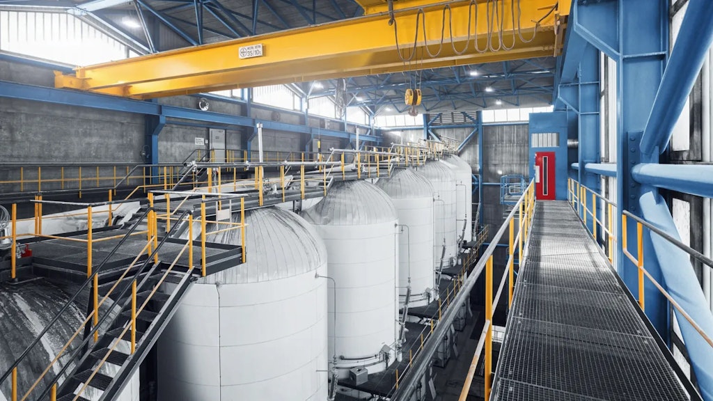 Vue intérieure d’un site industriel avec plusieurs grands réservoirs cylindriques, passerelles métalliques, garde-corps jaunes et pont roulant sous toiture.