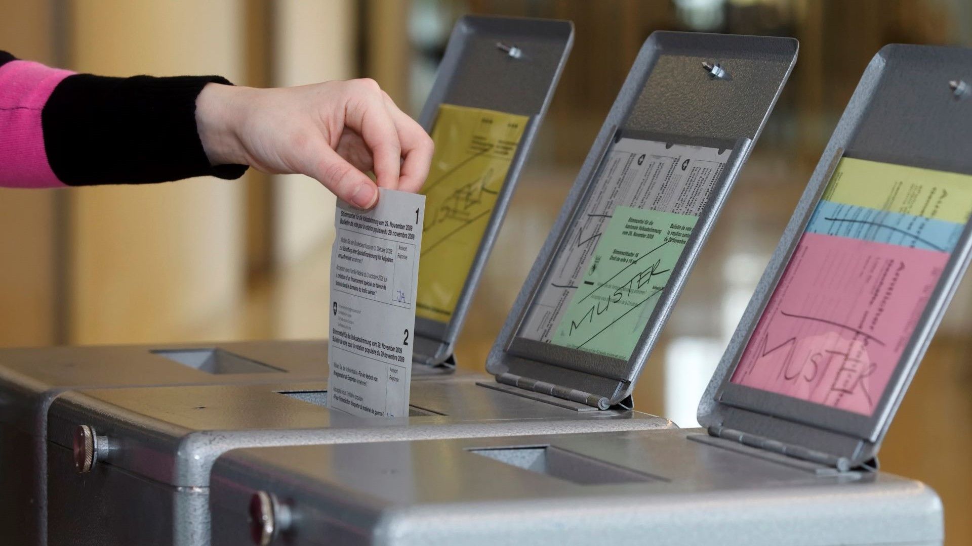 Une personne dépose un bulletin de vote dans une urne, tandis que plusieurs urnes contenant des bulletins de vote de différentes couleurs sont visibles.