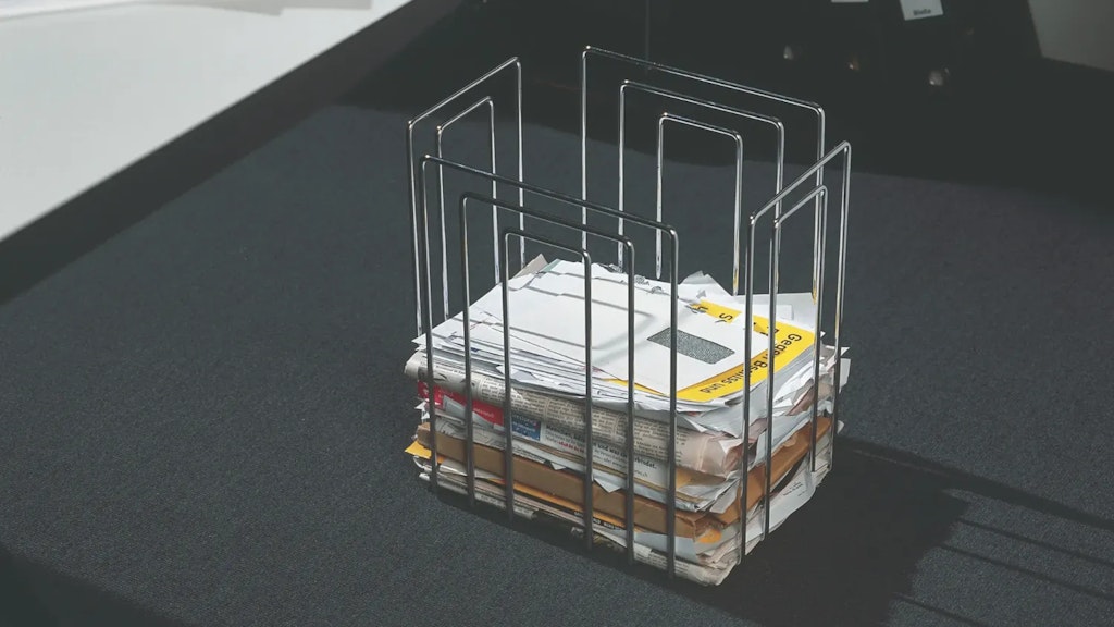 Porte-revues métallique contenant une pile de journaux et de courriers compressés, posé sur un sol sombre dans un intérieur.