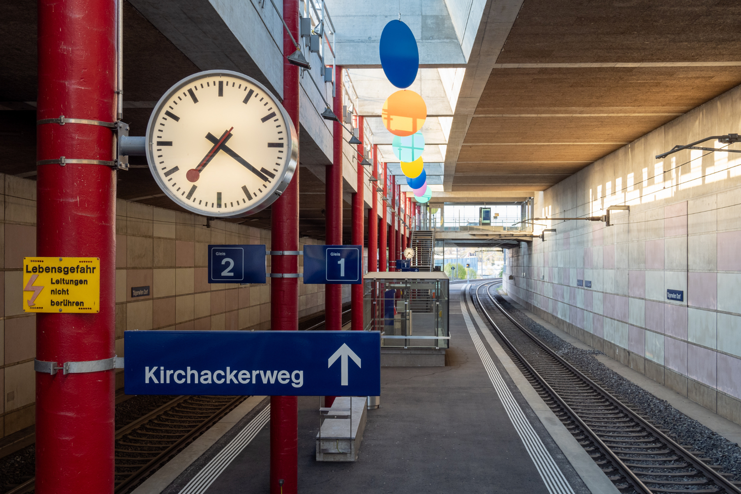 Une horloge murale, des panneaux de quai et des rails dans une gare souterraine avec des colonnes rouges et un panneau indiquant «Kirchackerweg».