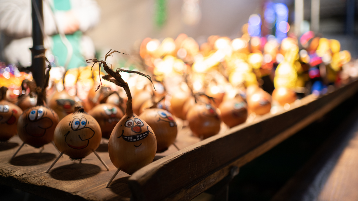 Oignons décorés avec des visages souriants exposés sur un étal de marché, éclairé, avec un vendeur indistinct en arrière-plan.
