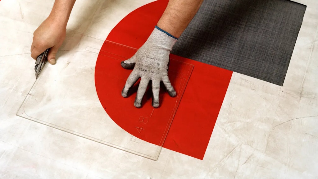 Mani che guidano una dima trasparente mentre tagliano un telone colorato con un cutter, con aree rosse, grigie e bianche visibili.