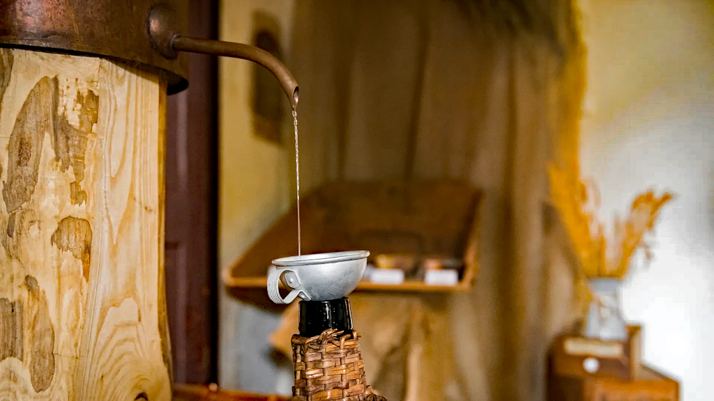 Liquido trasparente che scorre da un beccuccio in rame in un piccolo imbuto su una bottiglia, all’interno di una distilleria. 