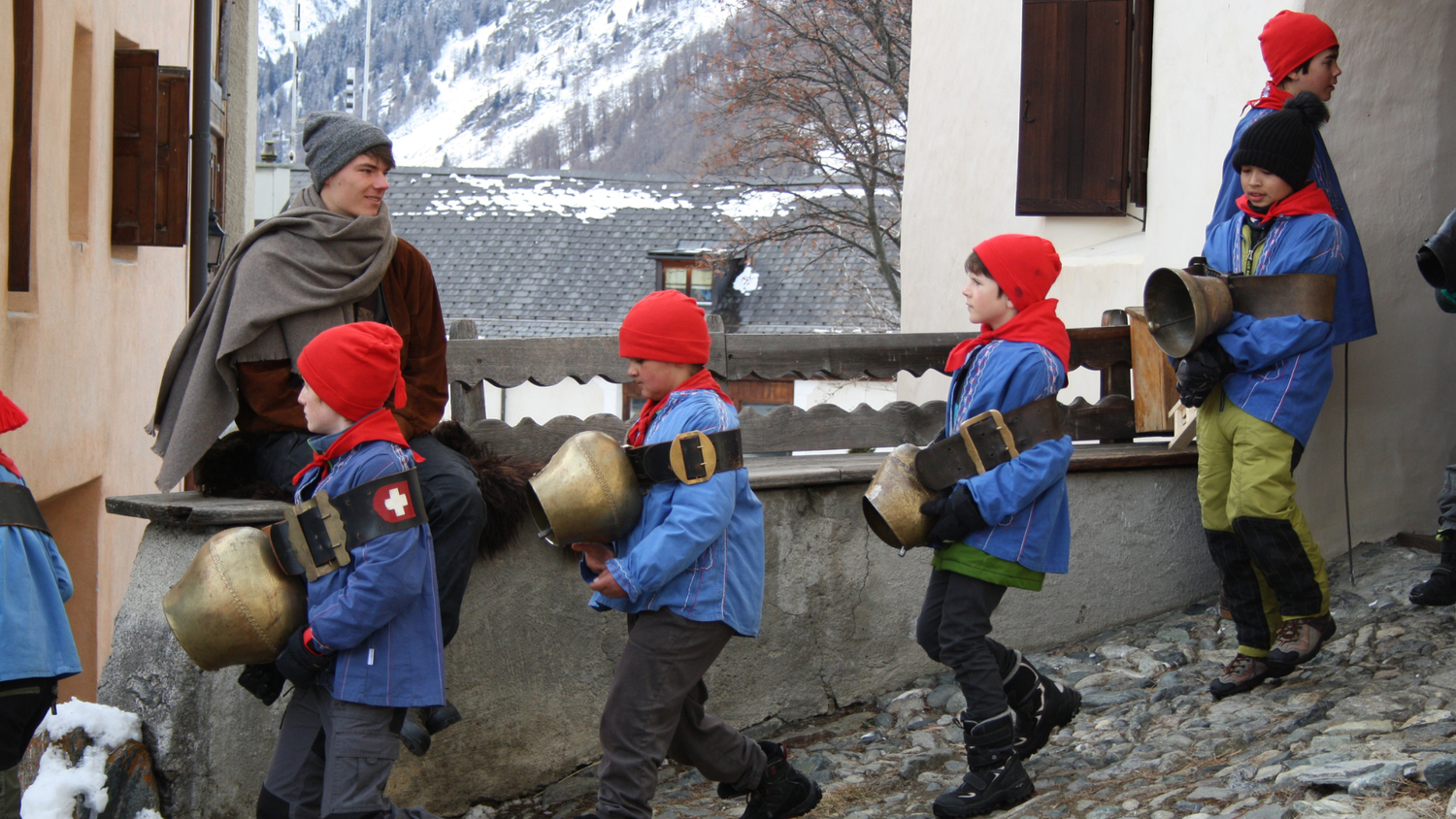 Bambini in abiti tradizionali blu con cappelli decorati e campane marciano in una strada innevata davanti a edifici alpini durante una processione.