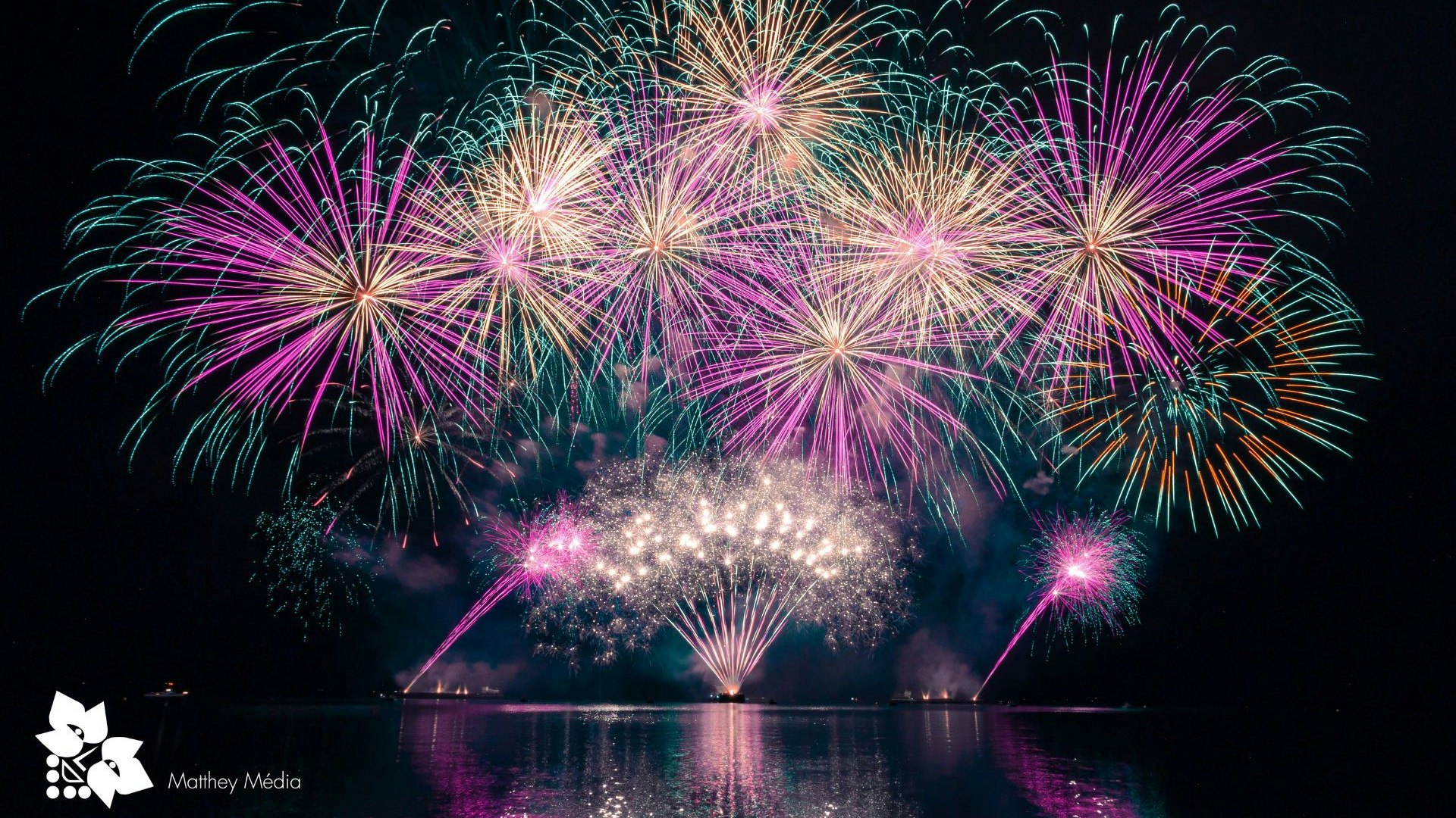 Fuochi d’artificio sparati da chiatte nella baia di Evole durante la festa della vendemmia, con riflessi colorati sull’acqua notturna.