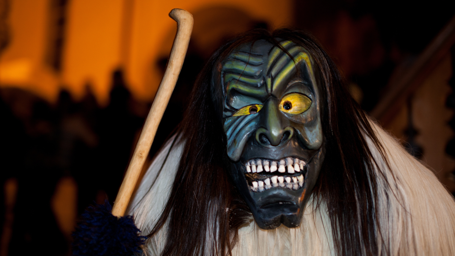 Persona mascherata con un volto grottesco dai tratti esagerati, lunghi capelli, un mantello di pelliccia e un bastone, durante un evento notturno.