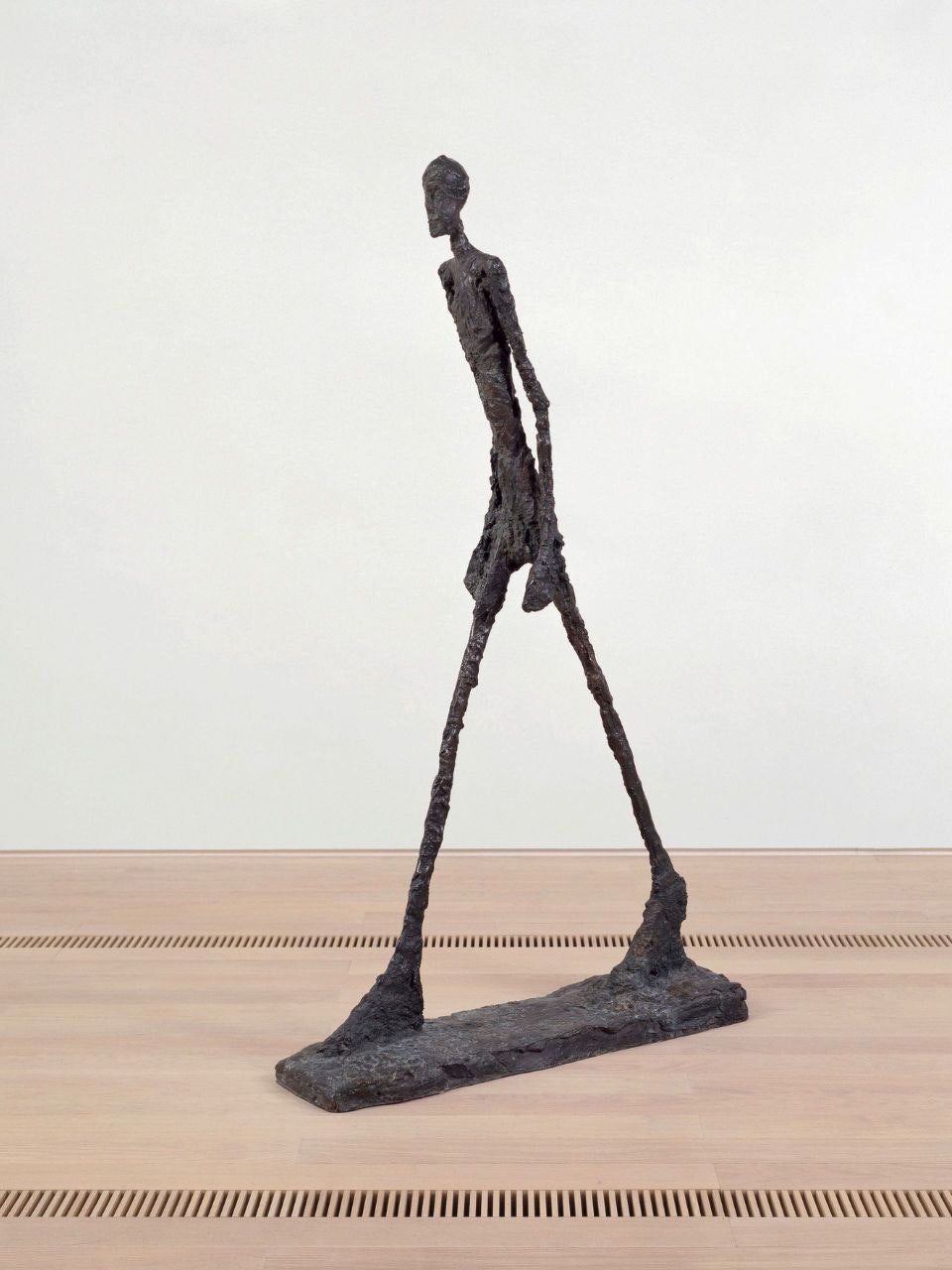 Fotografia de «L’homme qui marche II» (1960) di Alberto Giacometti: esile scultura in bronzo raffigurante una figura umana allungata in cammino, su basamento rettangolare. 