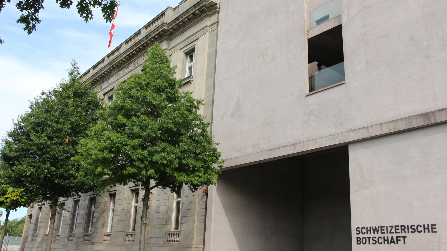 Facciata dell’ambasciata svizzera a Berlino, edificio moderno in cemento accanto a una struttura classica, con la bandiera svizzera sul tetto.