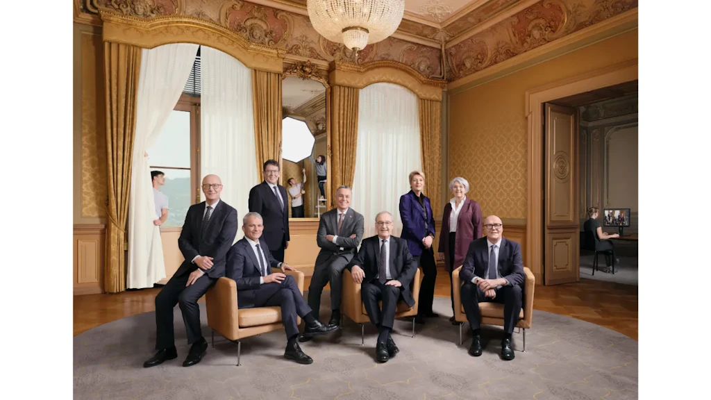 Foto del Consiglio federale svizzero in un salone. I suoi membri e il Cancelliere posano seduti o in piedi, circondati dal team tecnico e dalle attrezzature di studio.