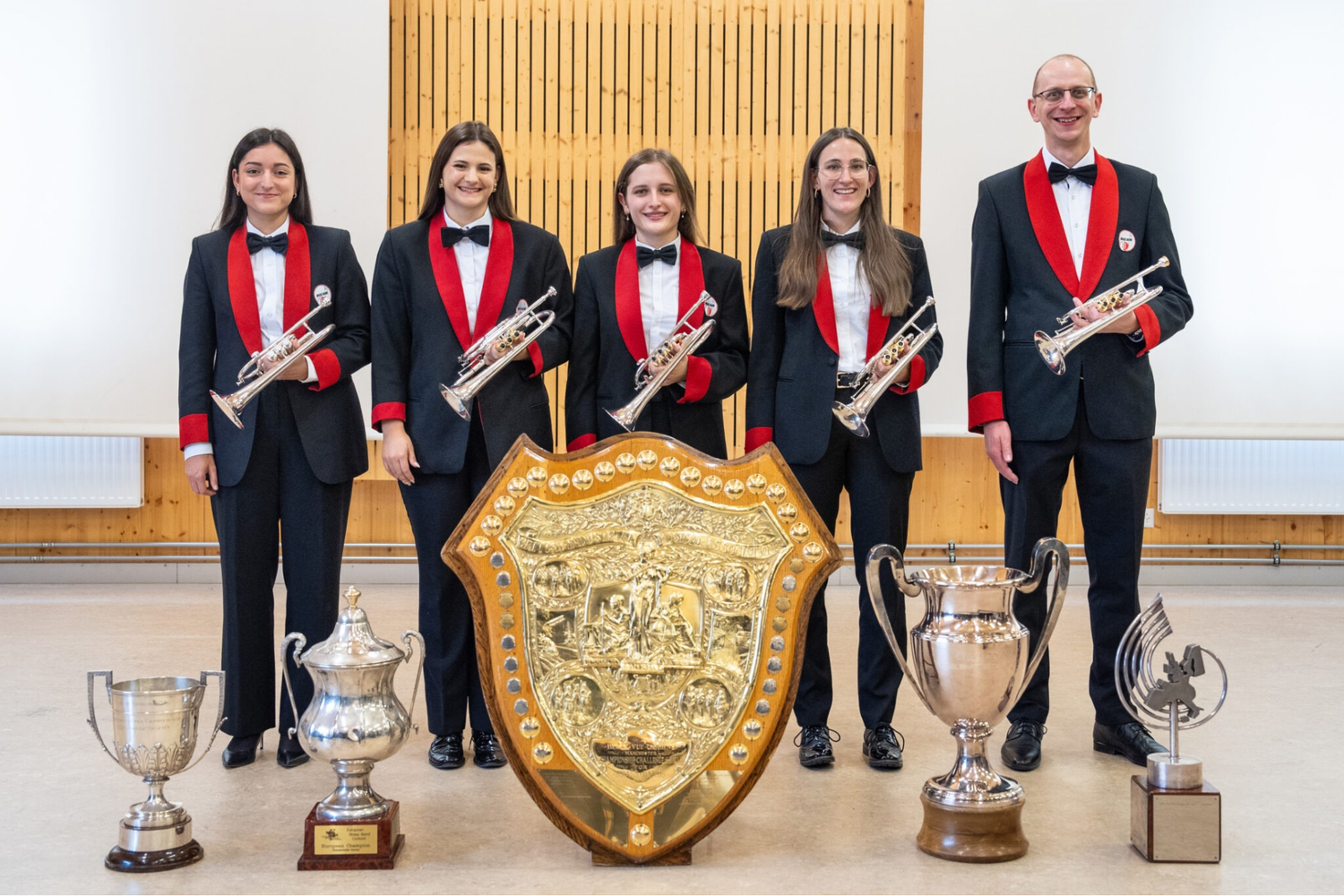 Cinque musicisti in frac nero con reverdi rossi tengono trombe, in piedi dietro cinque trofei in metallo e uno scudo dorato decorato, in una sala vuota.