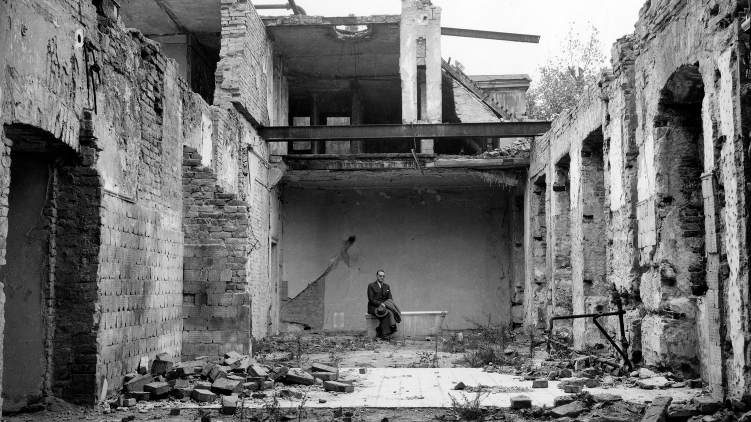 Foto in bianco e nero di un uomo in abito seduto su una vasca da bagno in un edificio in rovina, con pareti di mattoni danneggiate e detriti sul pavimento.
