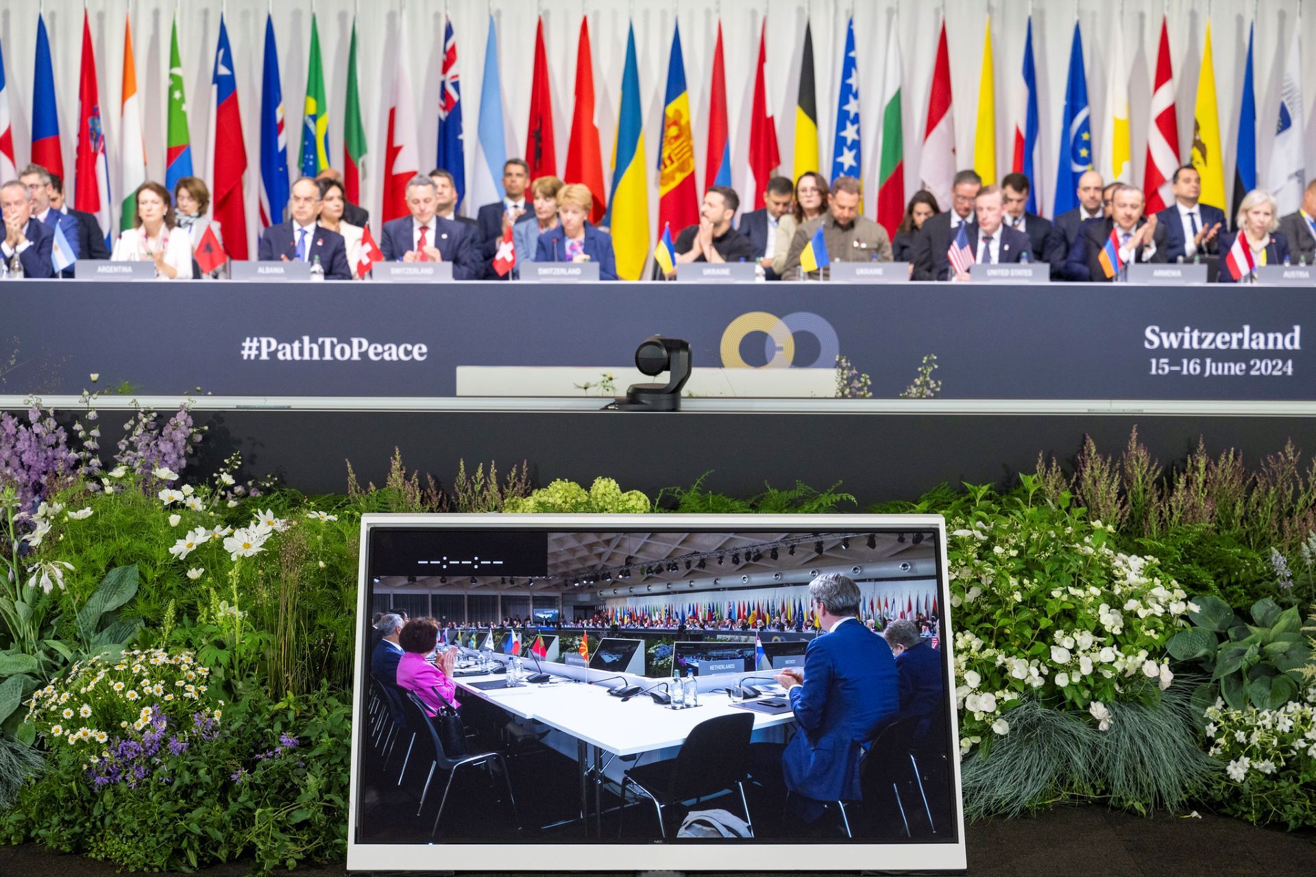 L’immagine mostra la conferenza internazionale #PathToPeace tenutasi in Svizzera nel 2024, con delegati di alto livello davanti alle bandiere nazionali e un monitor che mostra una videoconferenza.