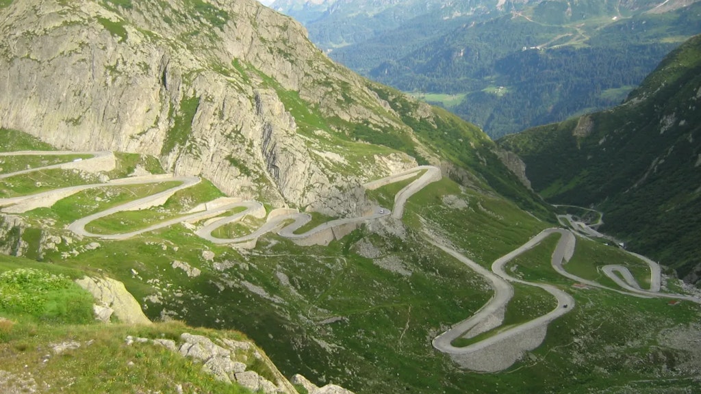 Strada di montagna tortuosa con molti tornanti che attraversa un paesaggio roccioso e verde, circondata da pendii ripidi e montagne sullo sfondo.