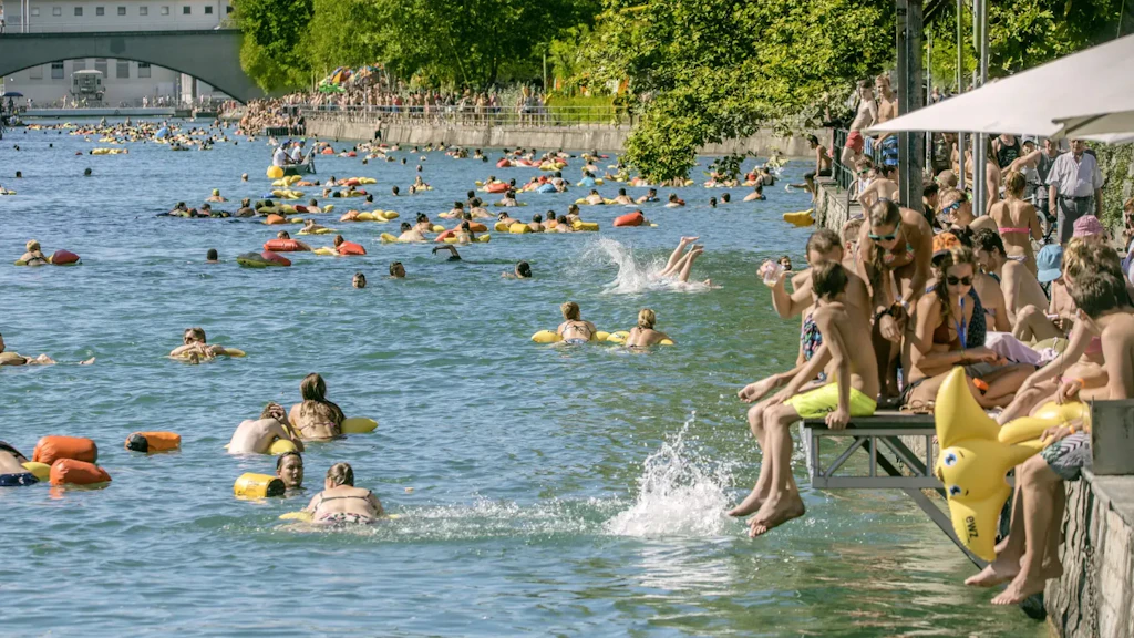 Numerose persone nuotano e galleggiano con salvagenti colorati in un fiume circondato da alberi in una giornata di sole.