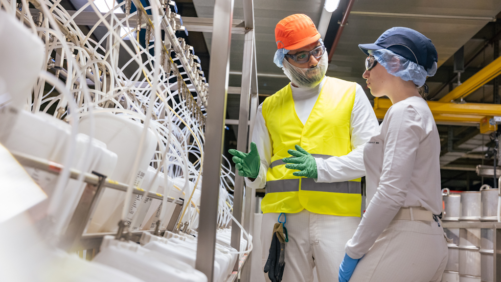 Due persone con abbigliamento protettivo parlano davanti a un impianto industriale con tubi e contenitori di plastica. 