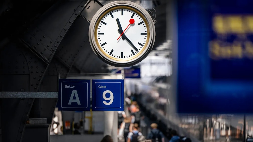Orologio ferroviario svizzero sopra i cartelli blu del settore A e del binario 9, con viaggiatori sfocati sul marciapiede.