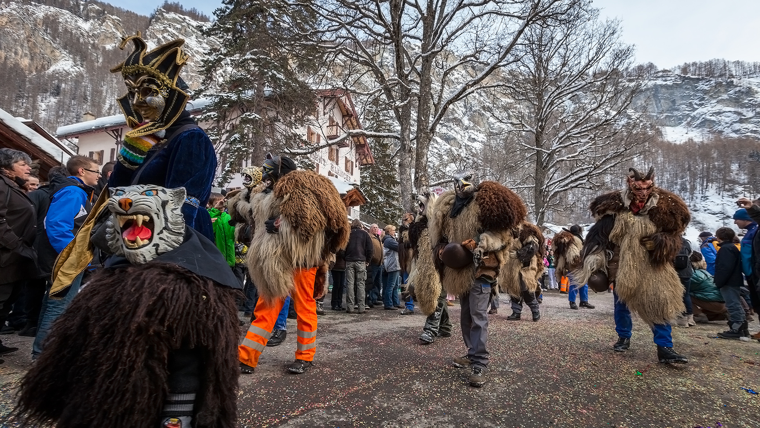 Persone in costumi di pelli animali con maschere scolpite a forma di teste di animalicamminano in una strada innevata, circondate da spettatori.