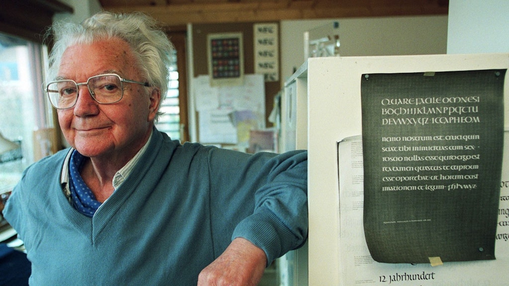 Un uomo anziano con maglione azzurro e occhiali appoggiato a un mobile su cui sono esposti esempi di calligrafia in un laboratorio luminoso. 