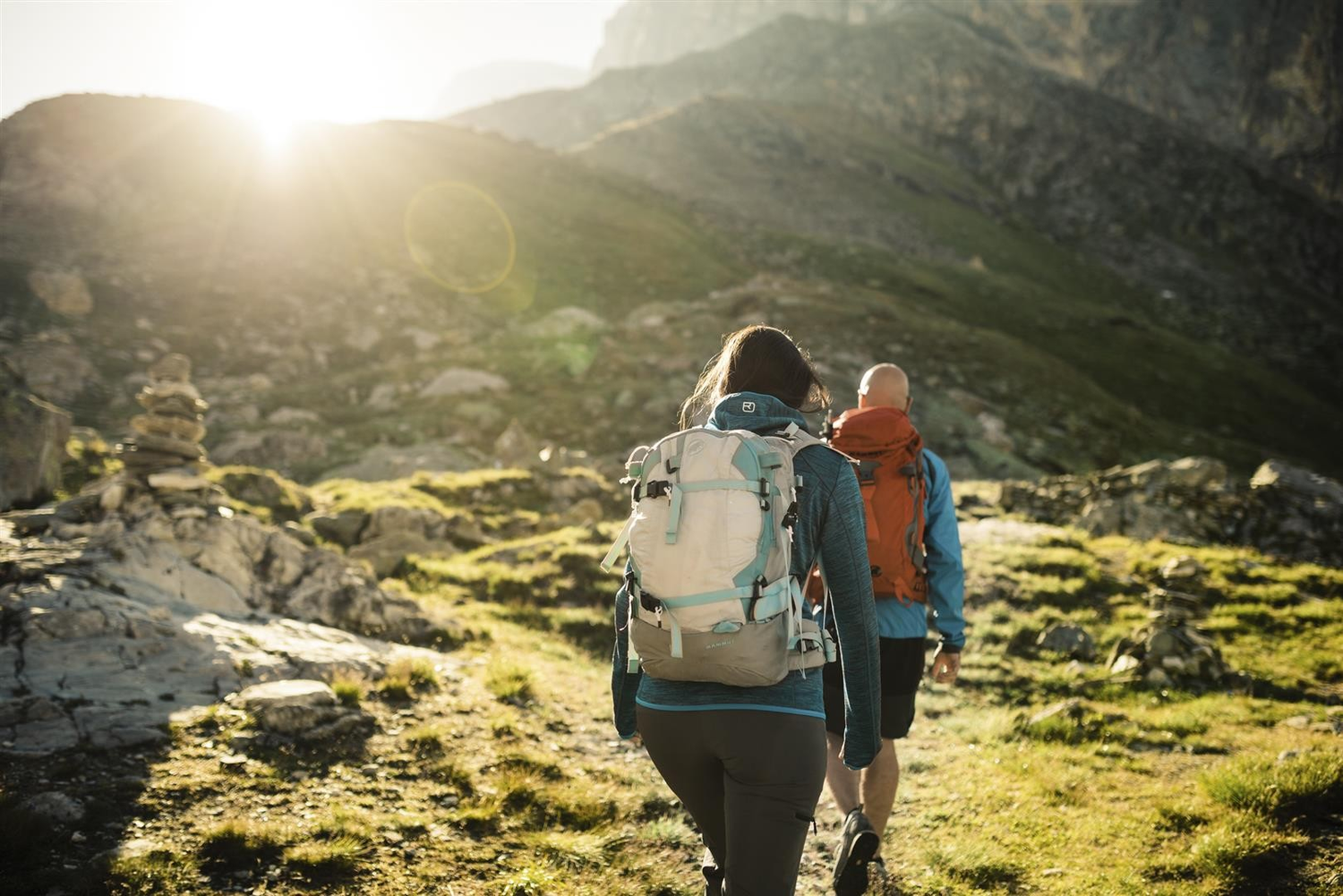 Due escursionisti camminano su un sentiero erboso in montagna, con il sole che splende luminoso sullo sfondo.