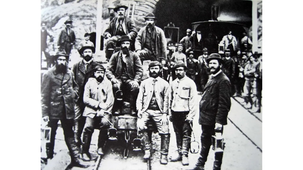 Gruppo di operai davanti all’ingresso di una galleria, con abiti da lavoro d’epoca, alcuni seduti su un carrello, altri in piedi accanto ai binari.