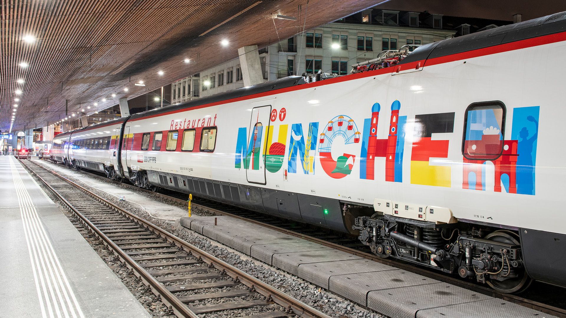 L’immagine mostra un treno in stazione con la scritta «Restaurant» e l’indicazione «MÜNCHEN» in caratteri colorati, decorato con i simboli della città di Monaco di Baviera.