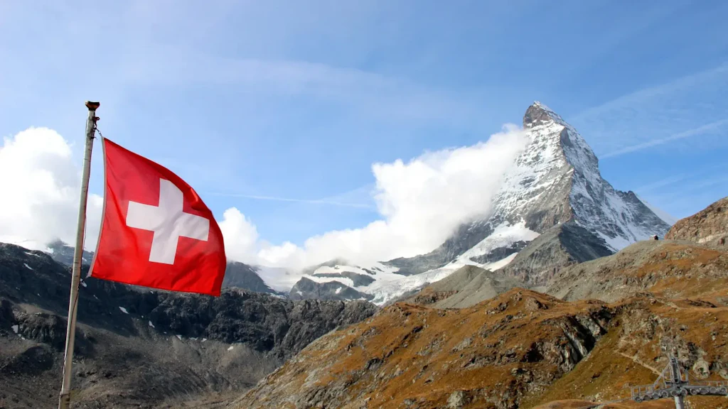 Bandiera svizzera rossa con croce bianca che sventola davanti a una montagna innevata sotto un cielo blu.