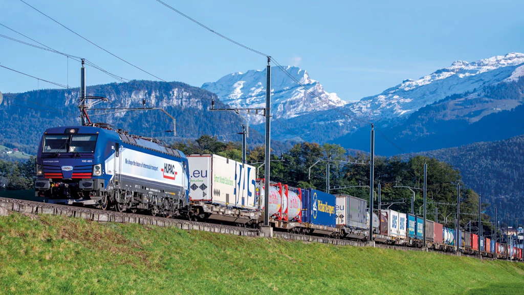 Un treno merci traina diversi vagoni container con vari tipi di merci attraverso un paesaggio montano.