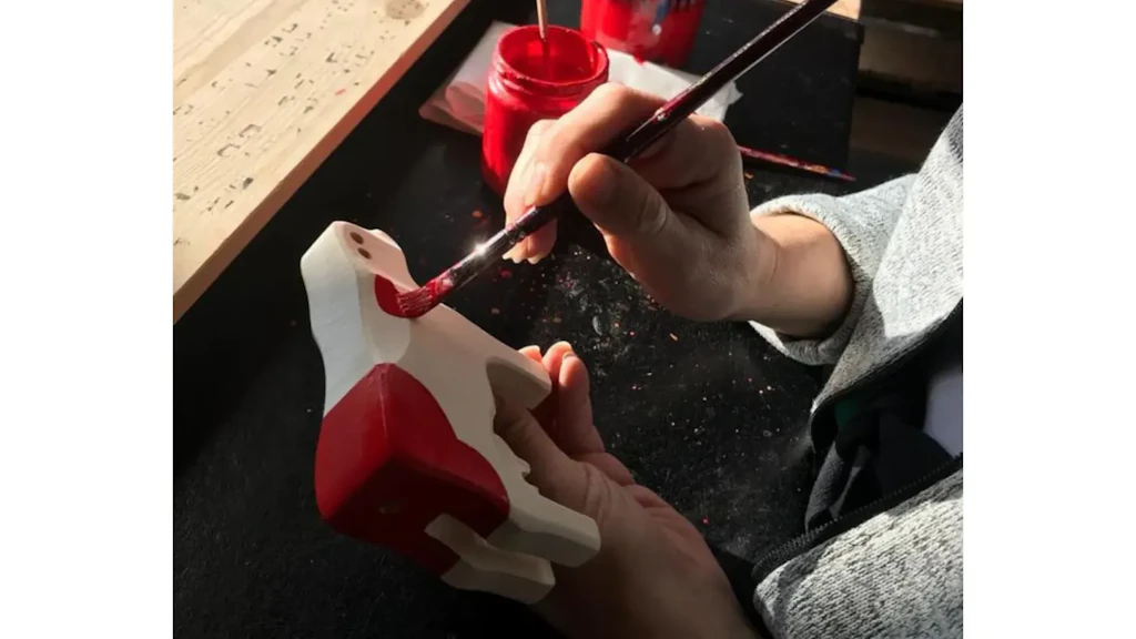 Una mano dipinge di rosso con un pennello una figurina in legno a forma di mucca, appoggiata su un piano di lavoro con vasetti di colore.