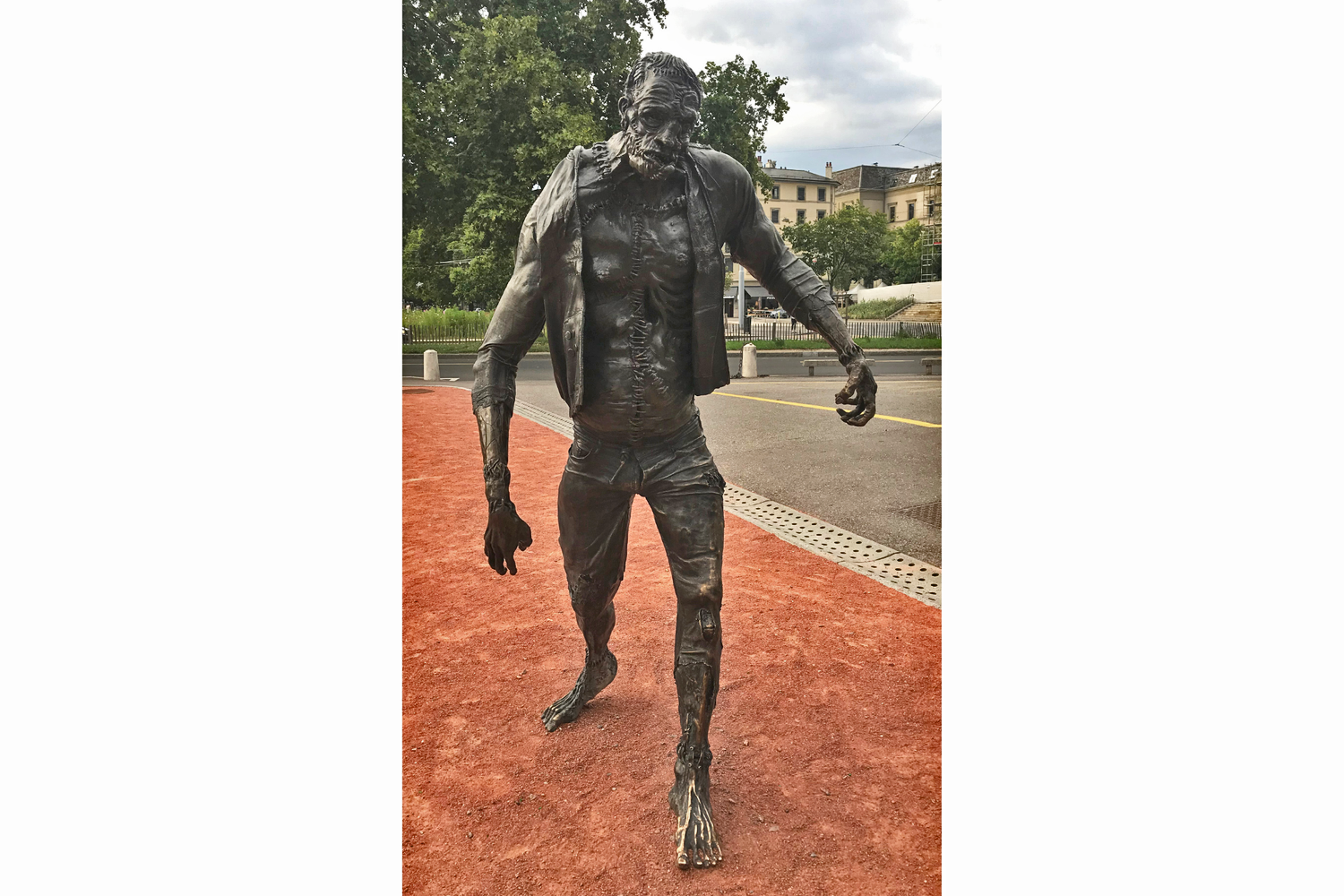 Scultura in bronzo di una creatura umanoide con arti asimmetrici, all’aperto su terreno rosso davanti a edifici urbani a Ginevra.