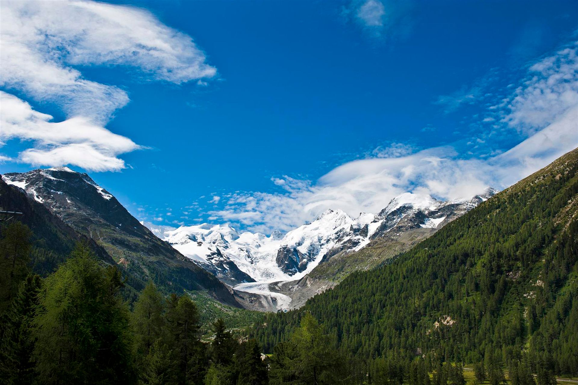 Vista de um vale alpino com florestas e picos nevados sob um céu azul com algumas nuvens.