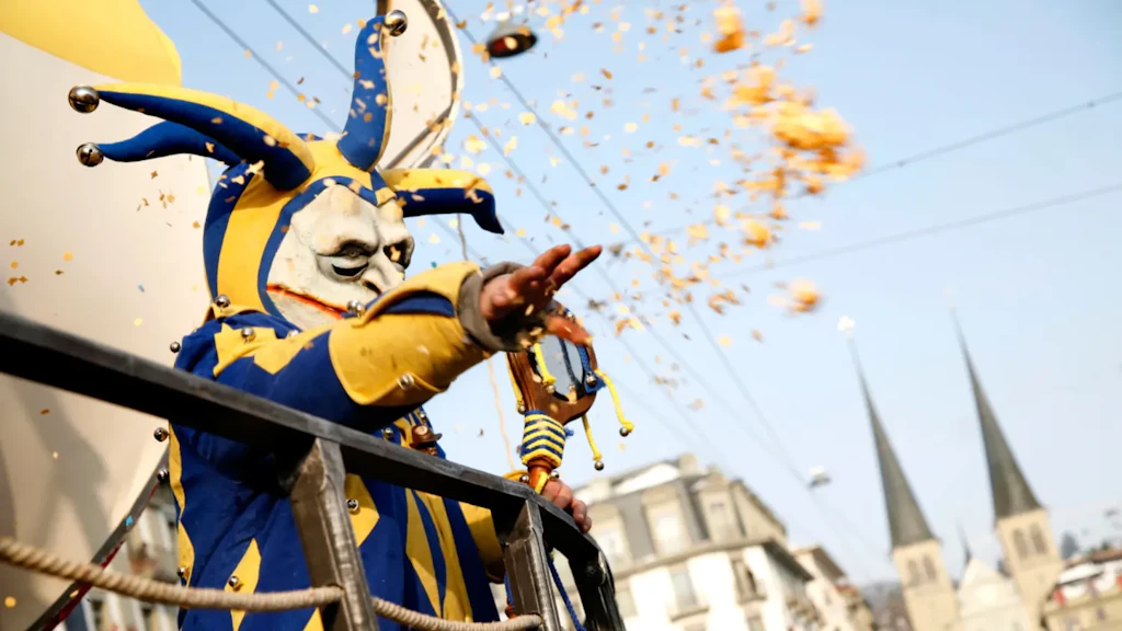 Personagem mascarado com fantasia azul e amarela lançando confetes de um carro alegórico, com prédios urbanos e torres de igreja ao fundo.