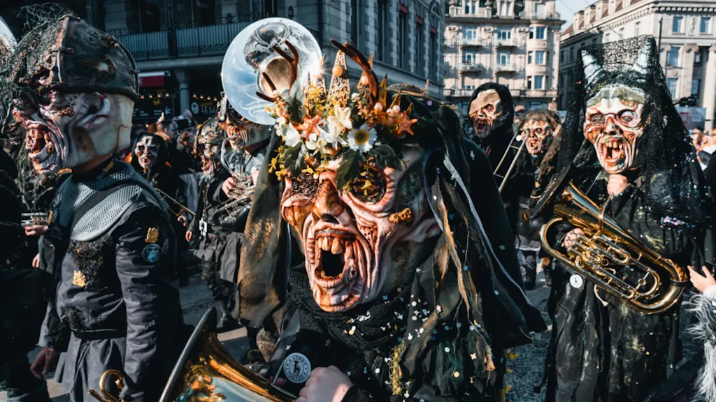 Grupo de pessoas com máscaras expressivas e trajes escuros em um desfile urbano, algumas segurando instrumentos de sopro metálicos.