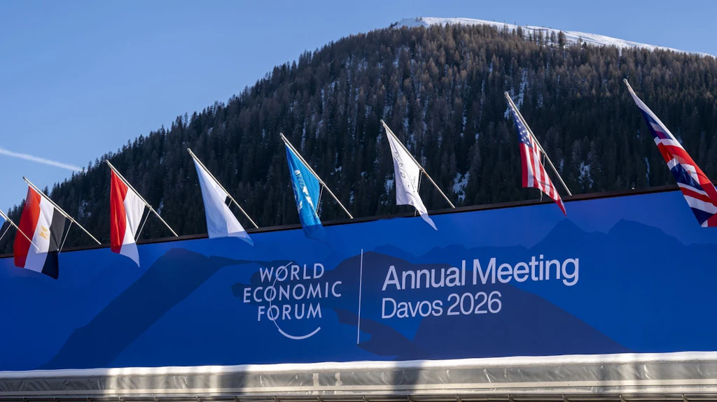 Diante de uma montanha coberta de neve, várias bandeiras nacionais tremulam acima de um painel azul com a inscrição «World Economic Forum – Annual Meeting». 